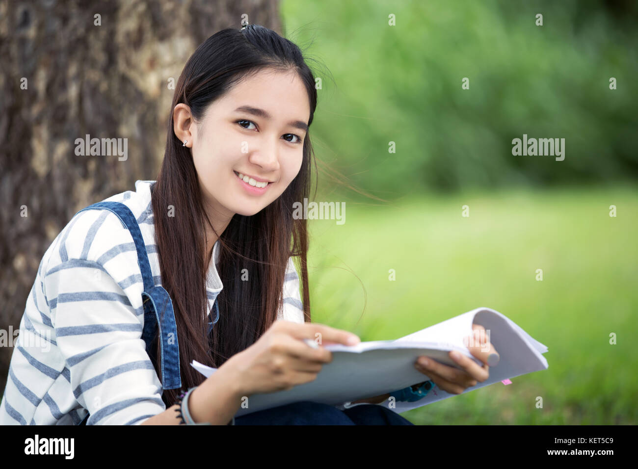 Smiling beautiful Asian girl reading book and working at tree on park ...