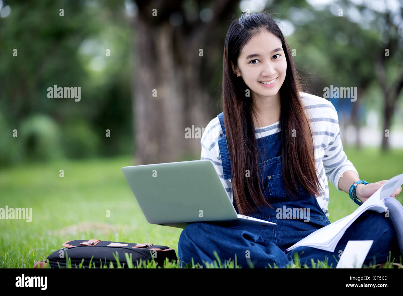 Smiling beautiful Asian girl reading book and working at tree on park ...
