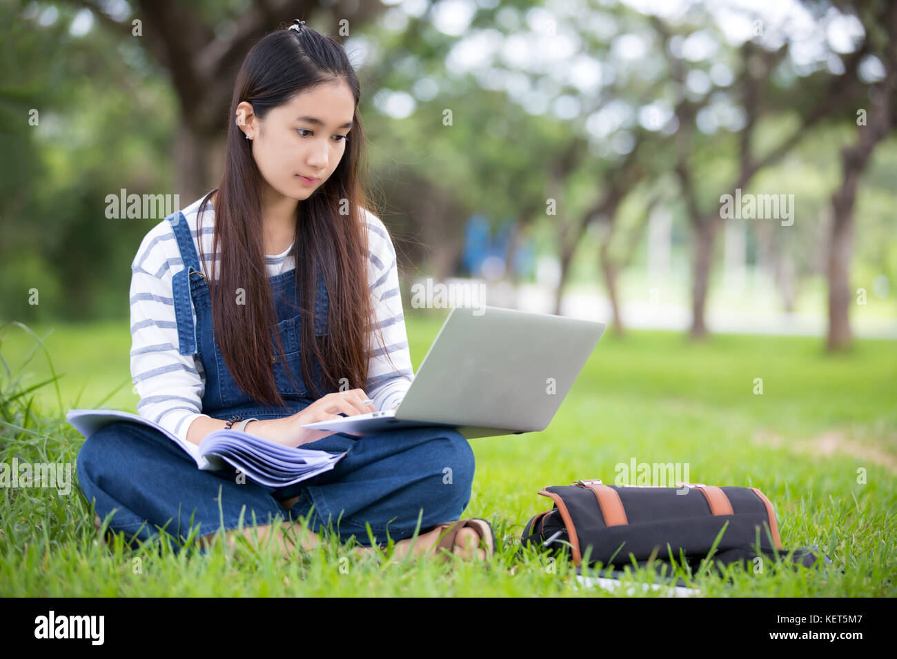 Smiling beautiful Asian girl reading book and working at tree on park ...