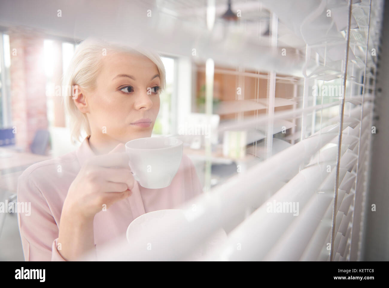 Businesswoman with coffee looking through blinds Stock Photo