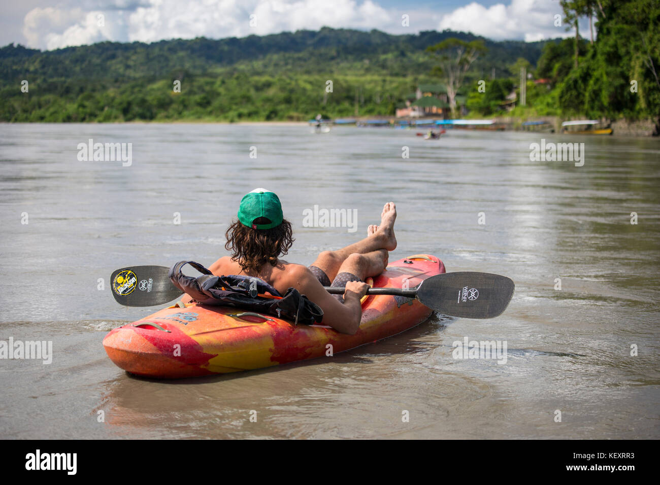 Young man relaxes in whitewater kayak on the Alto (Upper) Madre de Dios river int he jungles of Peru in the Peruvian Amazon. Village of Atalaya in the distance. Stock Photo