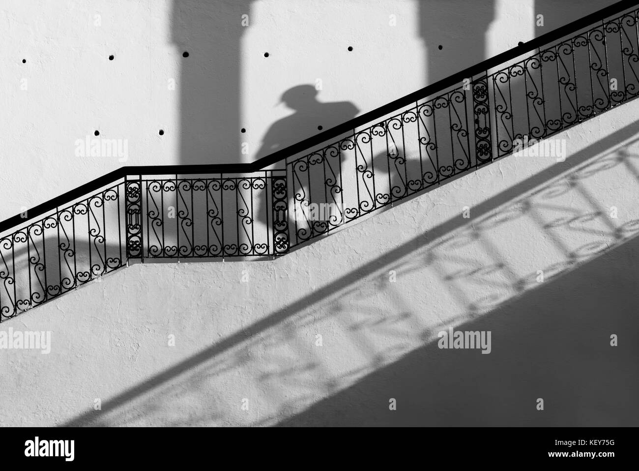 Staircase with black banister against white wall, with shadows of a man walking down. Black and white, monochrome image. Stock Photo