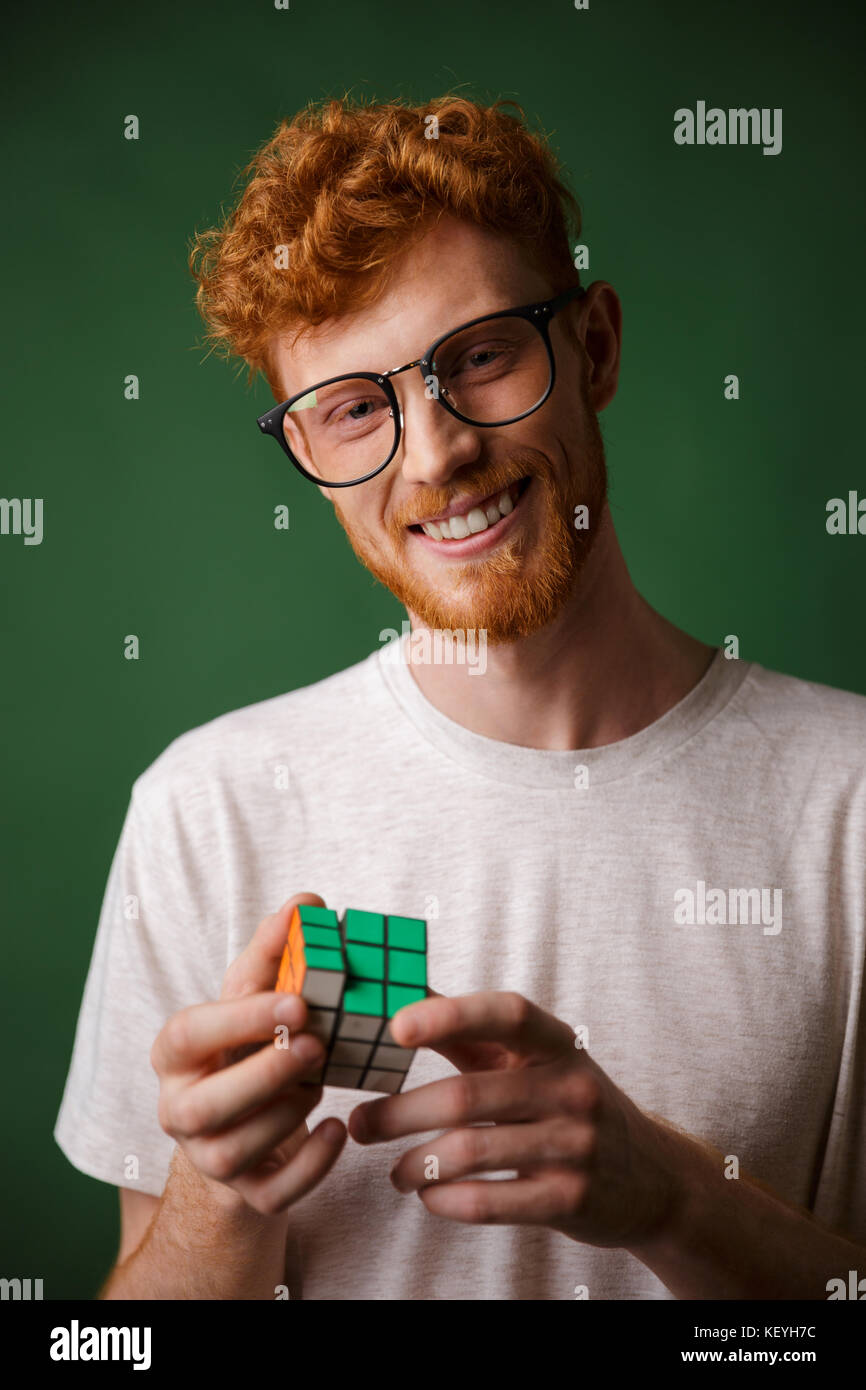 Young smart readgead bearded man in glasses, playing with rubic's cube, looking at camera, over green background Stock Photo