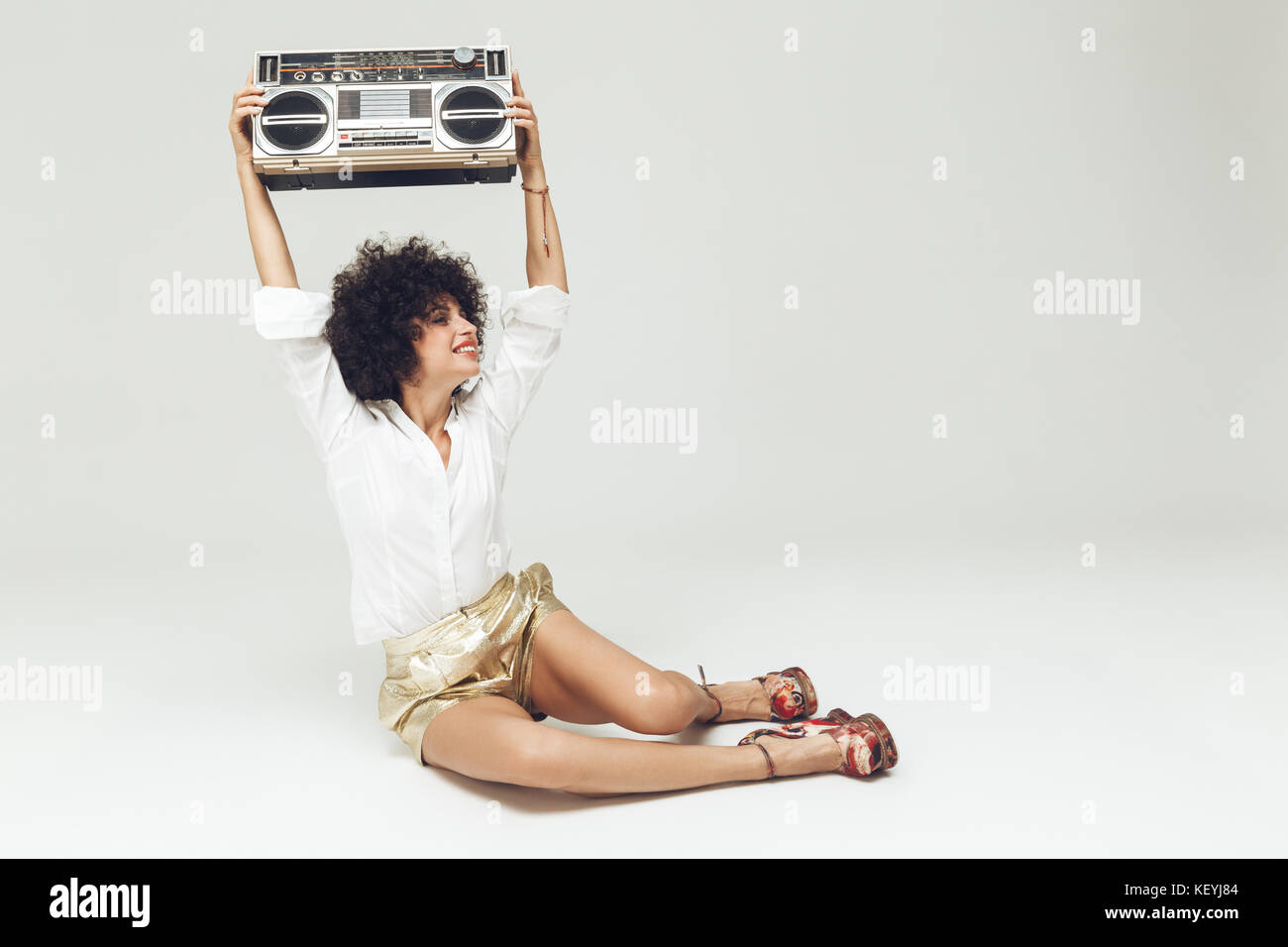 Image of young beautiful retro woman dressed in shirt sitting and posing isolated. Looking aside near boombox. Stock Photo