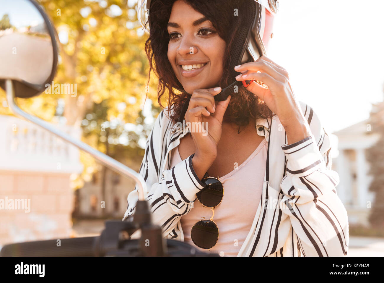 Close up image of happy african woman sitting on modern motorbike and fastens her moto halmet outdoors Stock Photo