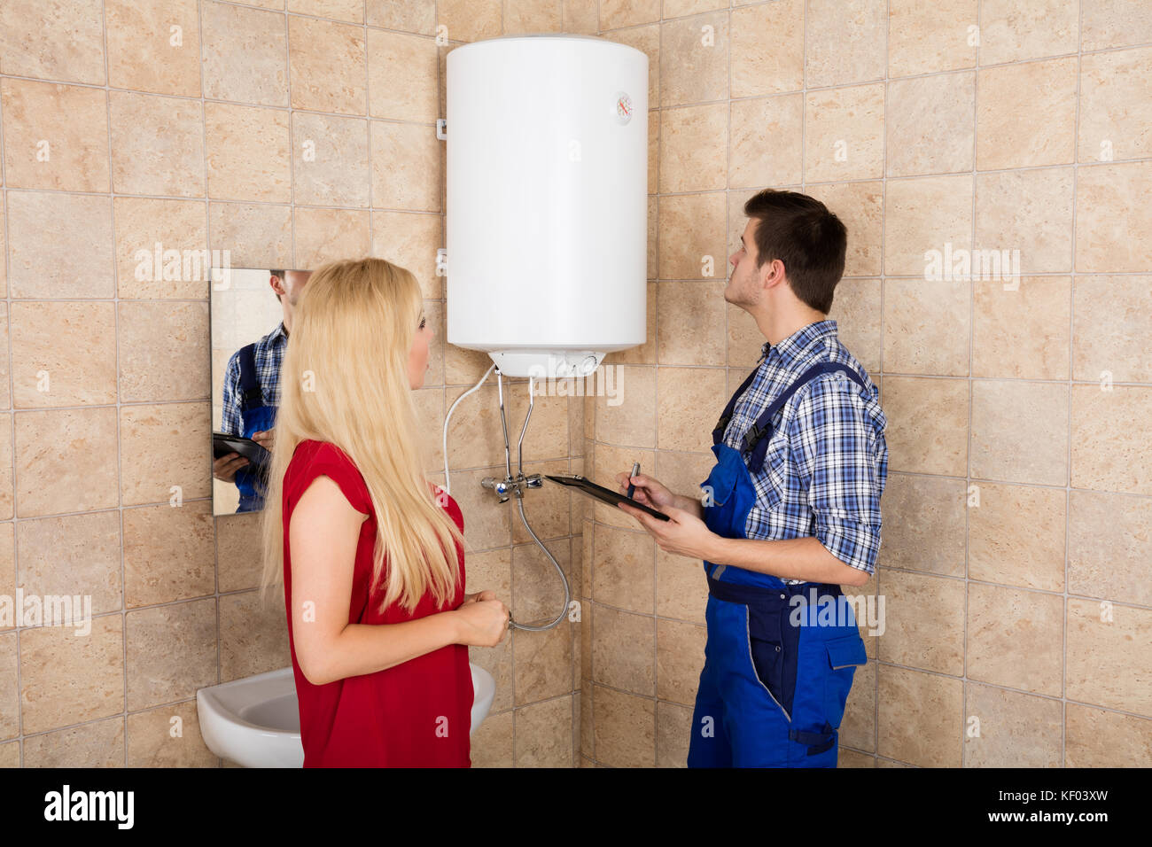Woman Looking At Young Male Plumber Holding Clipboard Looking At Electric Boiler Stock Photo