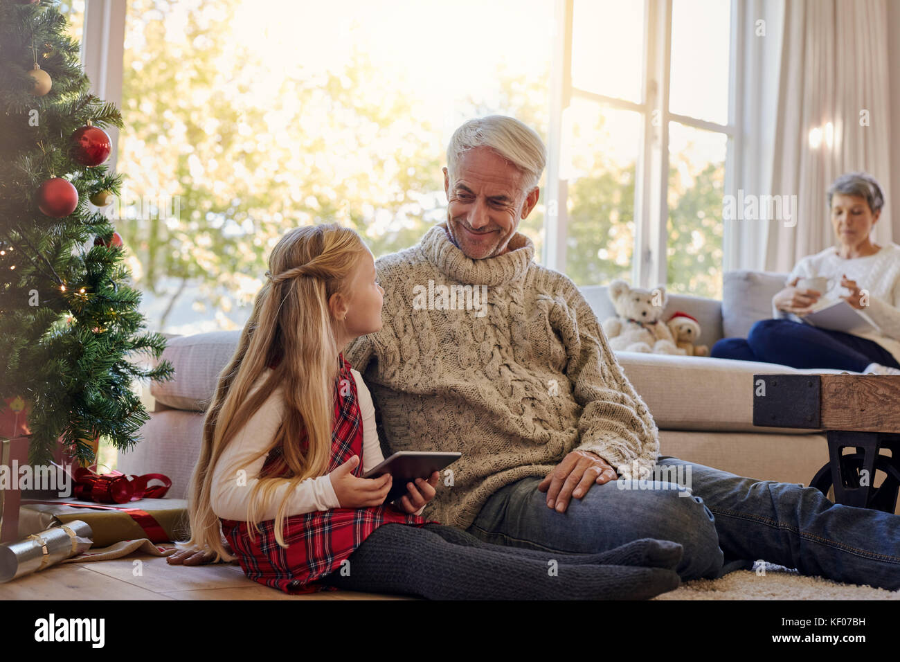 Little girl and grandfather sitting on floor by christmas tree with digital tablet, with grand mother sitting on sofa at the back. Stock Photo
