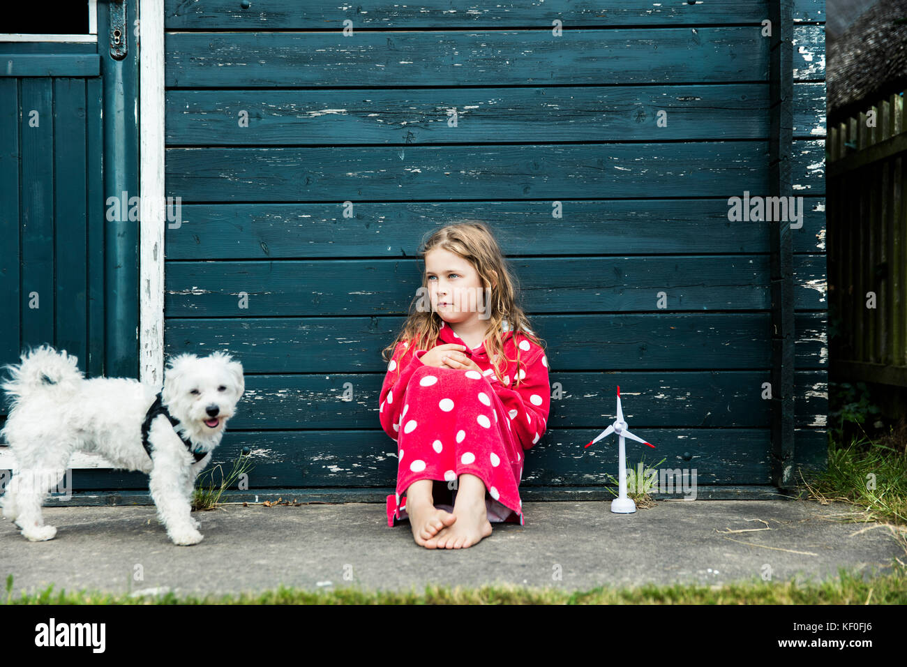Girl wearing bathrobe outdoors sitting at wooden hut with toy wind turbine and dog Stock Photo