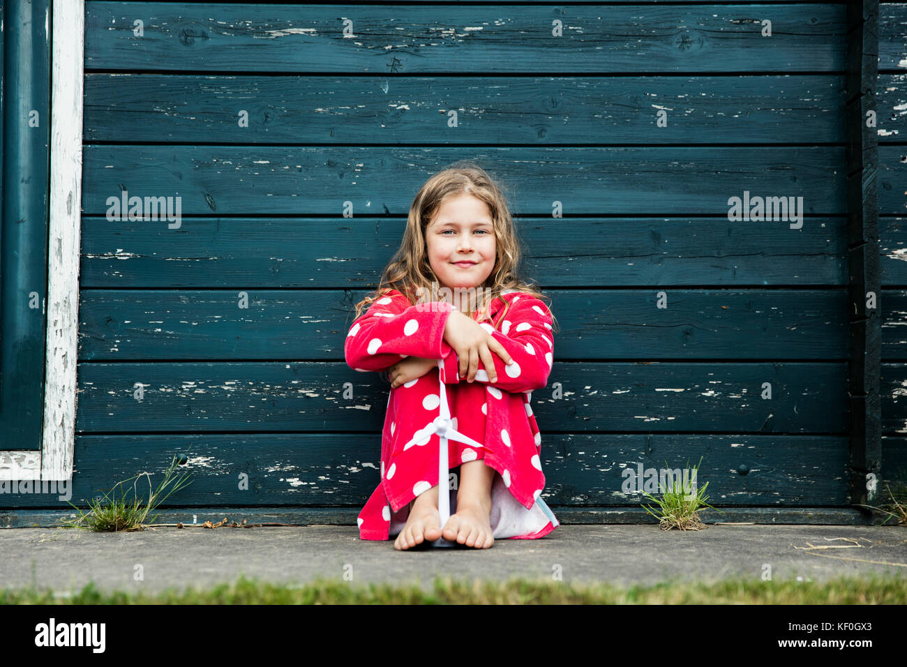 Girl wearing bathrobe outdoors sitting at wooden hut with toy wind turbine Stock Photo