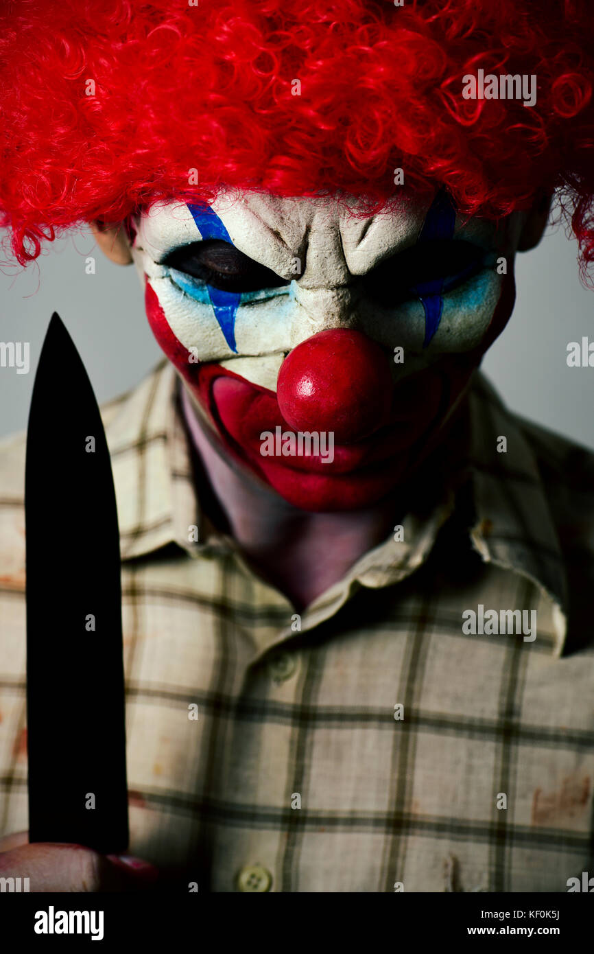 closeup of a scary evil clown wearing a dirty and ragged plaid shirt with stains of blood, looking down while holding a big knife in his hand Stock Photo