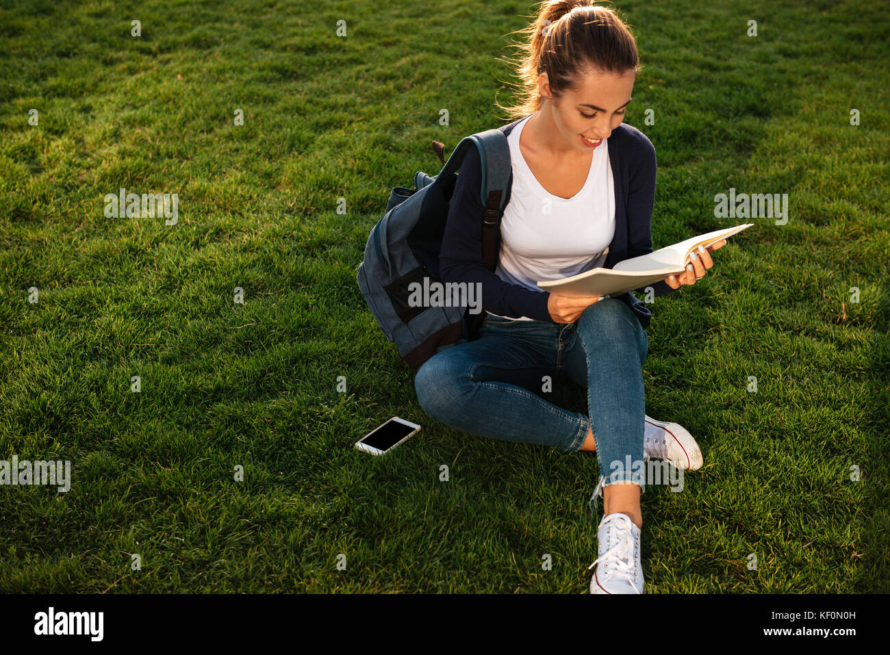 Portrait of a casual young student girl with backpack reading textbook ...