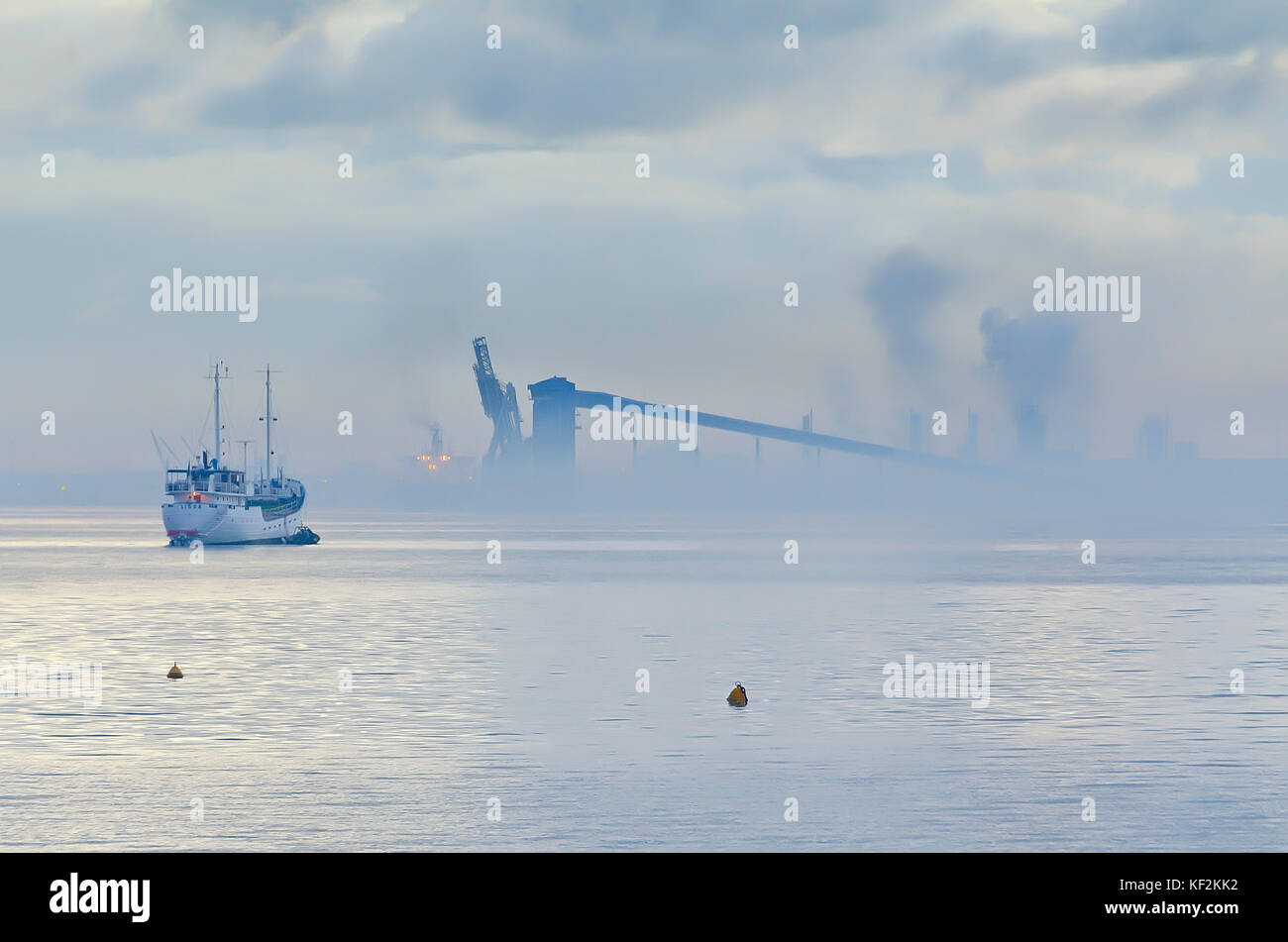 White ship at anchor in quiet harbour on misty morning with industry in background Stock Photo