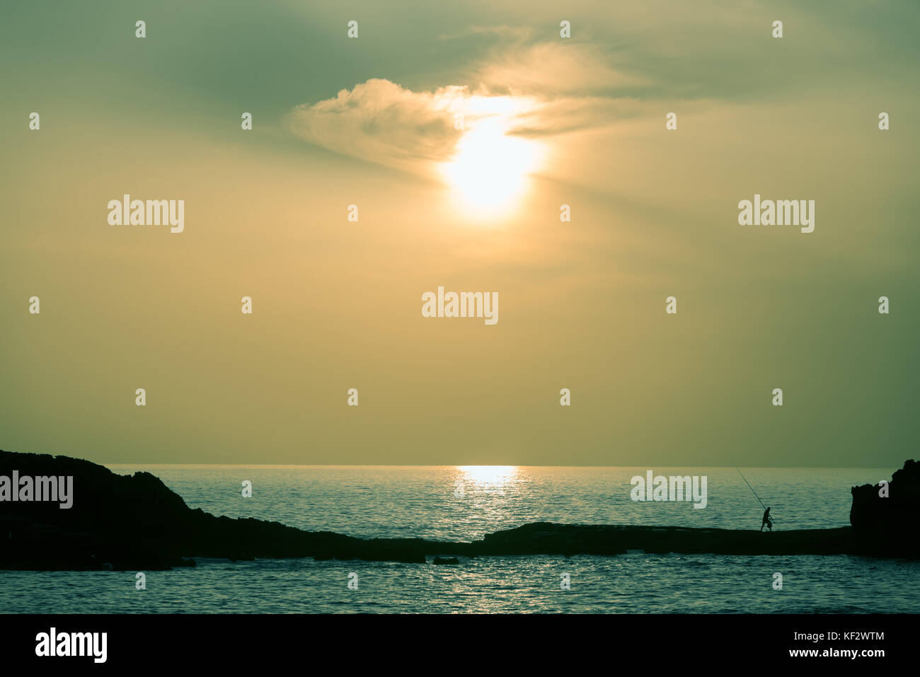 Silhouette of a fisherman walking on a rock at the sea at sunset. Stock Photo