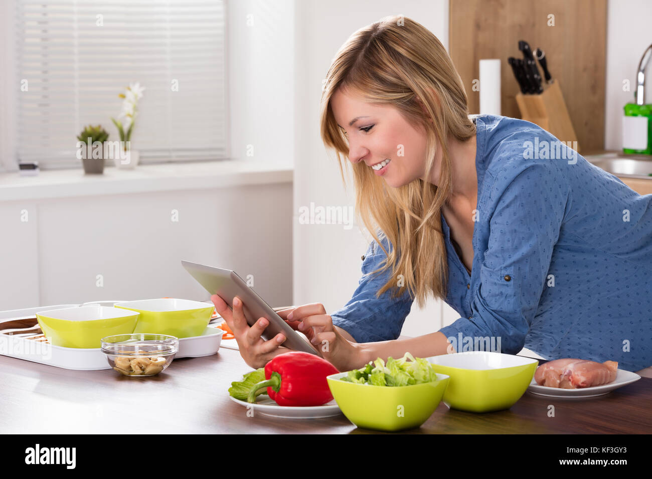 Smiling Young Woman Using Digital Tablet With Vegetable On Countertop In Kitchen Stock Photo