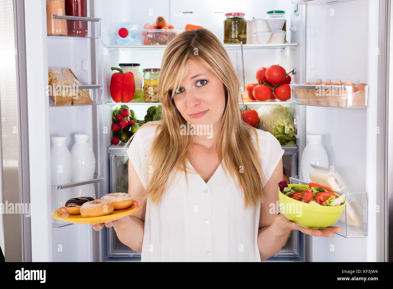 Confused Young Woman Holding Salad And Plate Of Donut Near Refrigerator Stock Photo