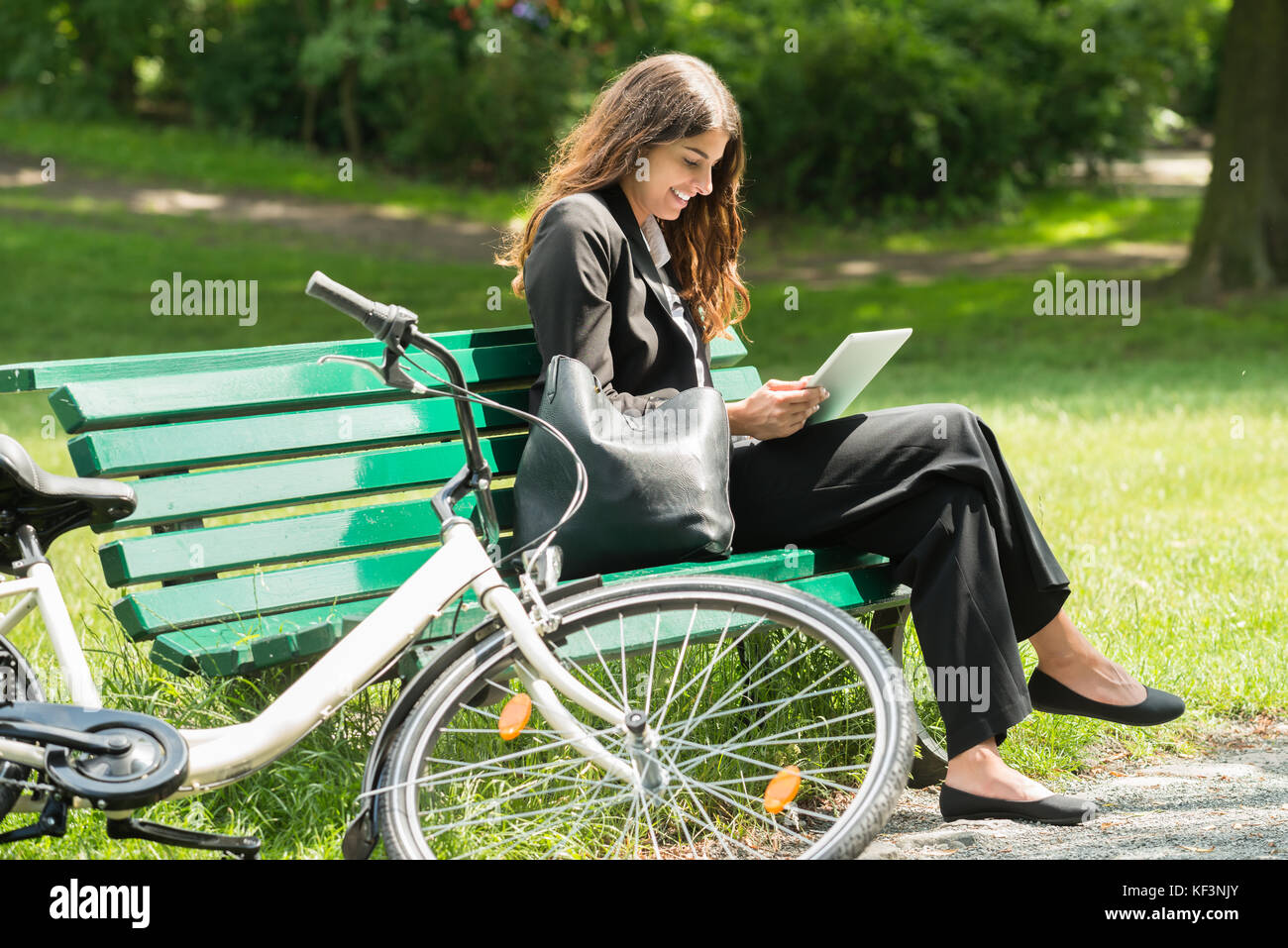 Smiling Young Businesswoman Sitting On Bench Using Digital Tablet At Park Stock Photo