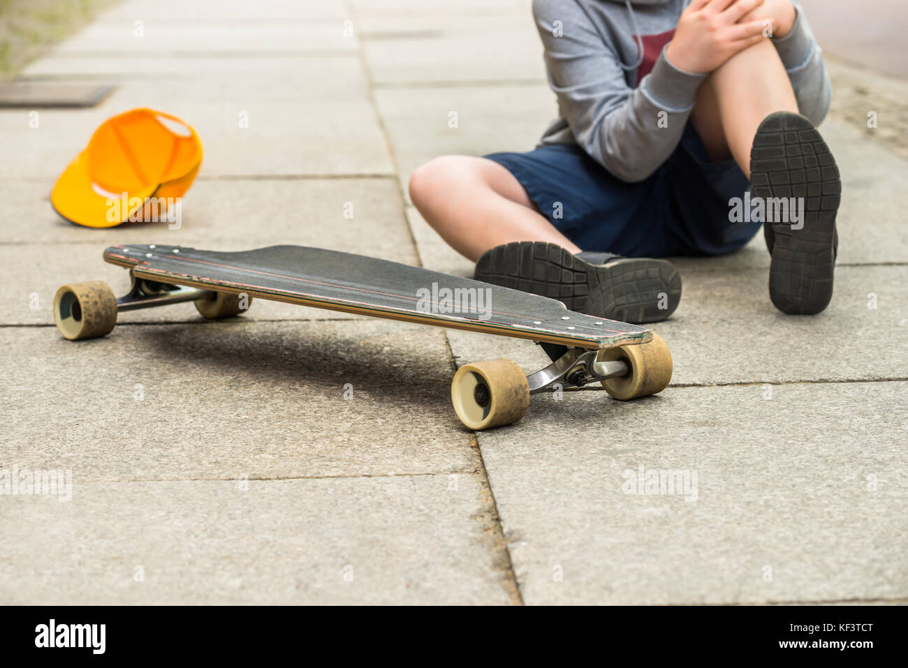 Boy With Knee Injury Sitting Near Skateboard On Sidewalk Stock Photo