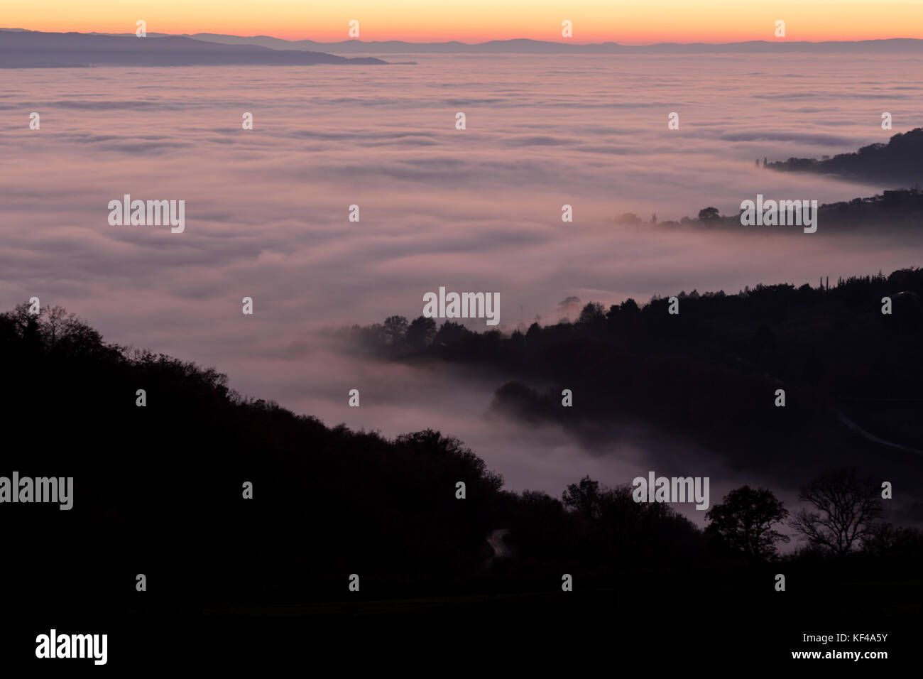 Aerial view of a valley filled by fog at sunset, with beautiful warm colors of the sky reflecting on the mist Stock Photo