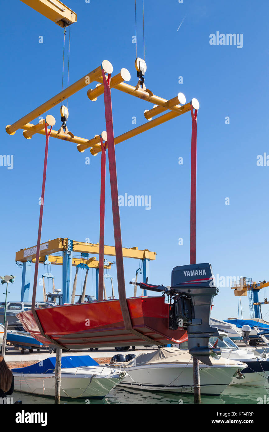 Small red boat with outboard motor suspended from a boat lift in a sling over water  at a marina during launch Stock Photo