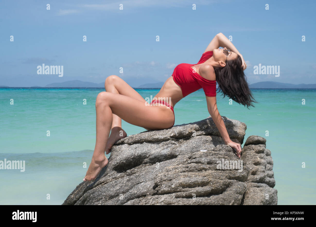 Beautiful brunette woman in red bikini and sunglasses posing on the rock of sandy paradise island beach over blue sea and sky background Stock Photo