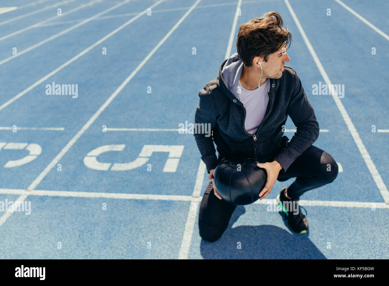 Athlete sitting on a running track near the start line with a medicine ball looking away. Runner wearing earphones sitting on the running track holdin Stock Photo