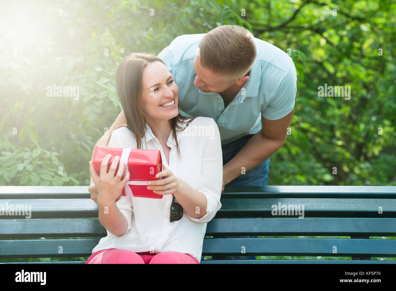 Young Man Giving His Wife A Surprise Gift While Sitting On Bench In Park Stock Photo
