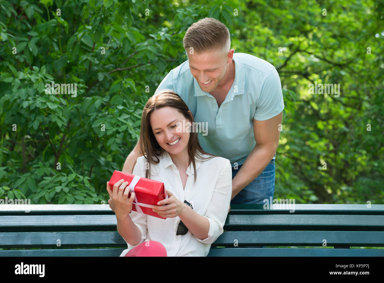 Young Man Giving His Wife A Surprise Gift While Sitting On Bench In Park Stock Photo