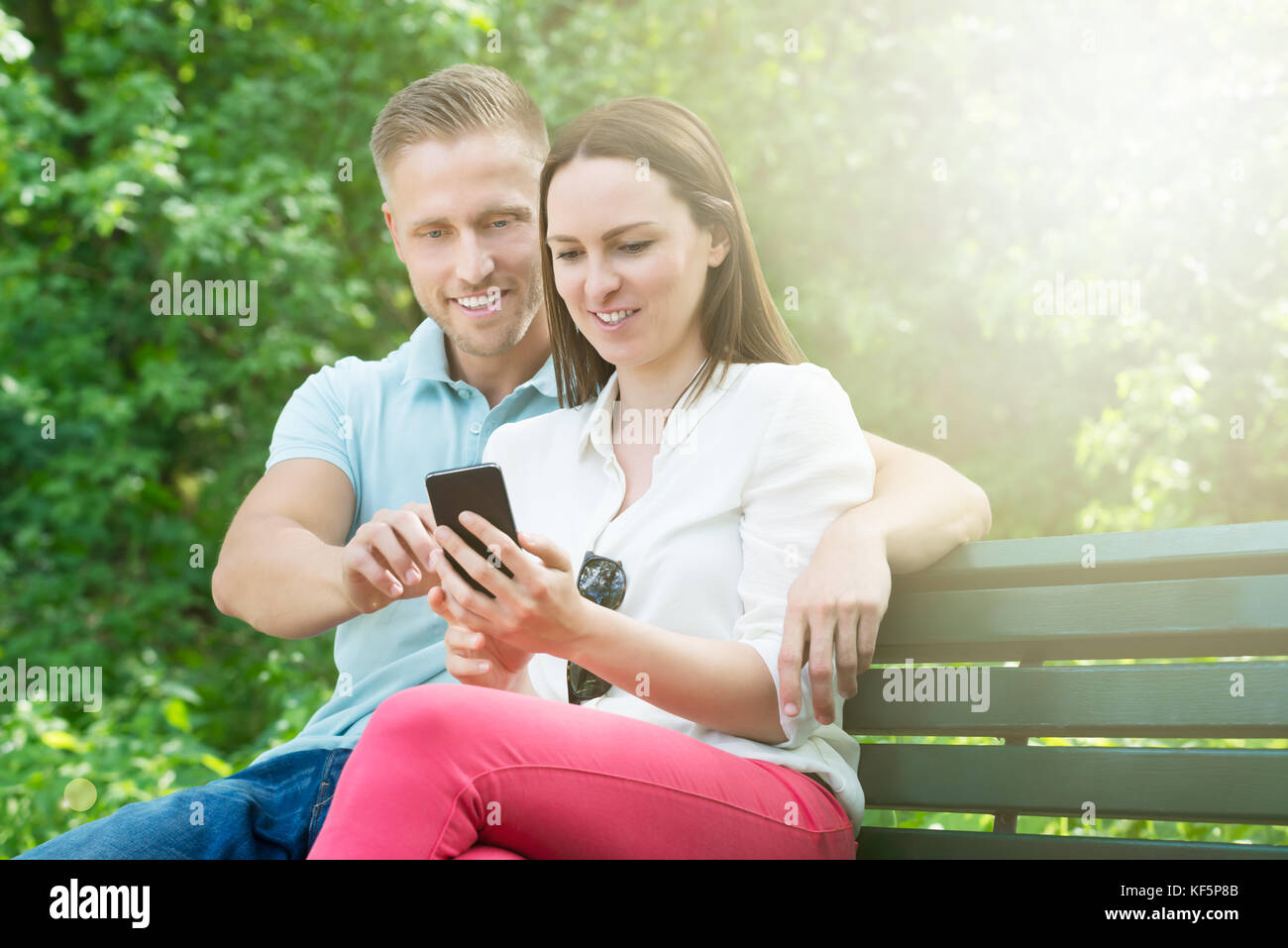 Young Happy Couple Using Mobile Phone While Sitting On Bench In Park Stock Photo