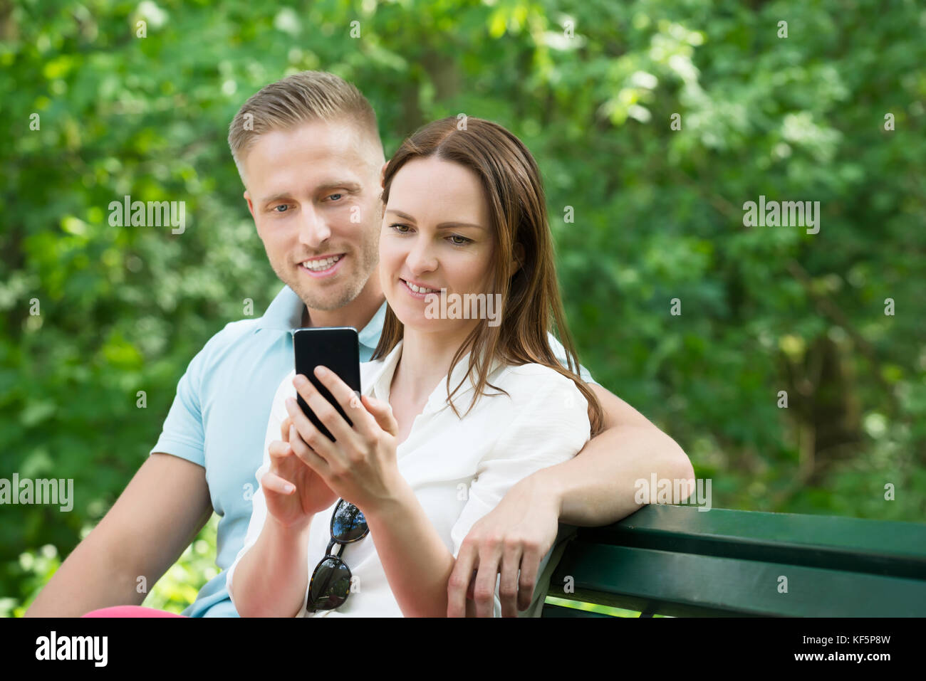 Young Happy Couple Using Mobile Phone While Sitting On Bench In Park Stock Photo