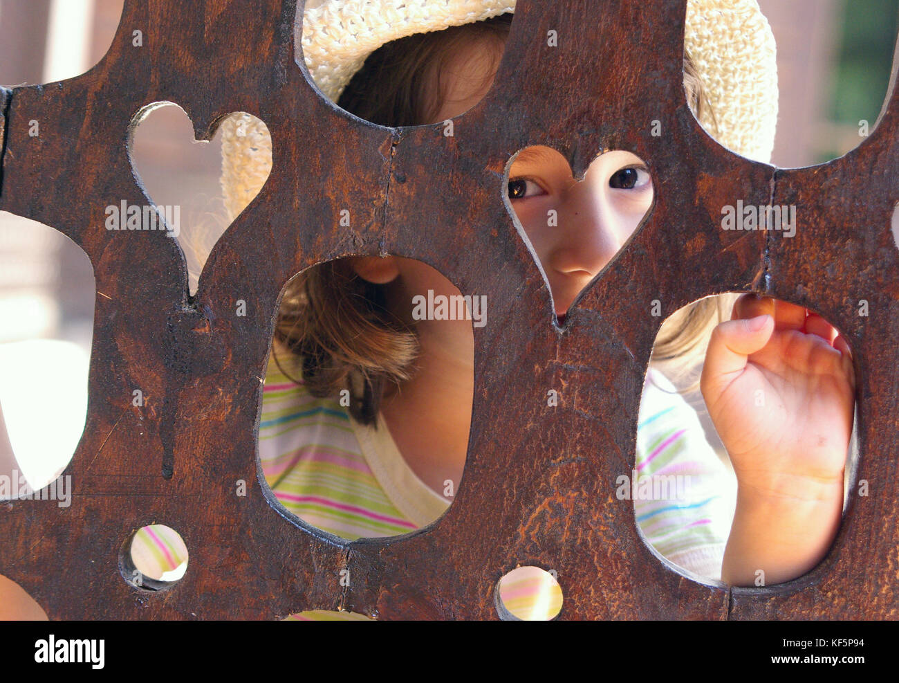 Cute little girl peeking behind heart shaped wooden fence, looking with curiosity Stock Photo