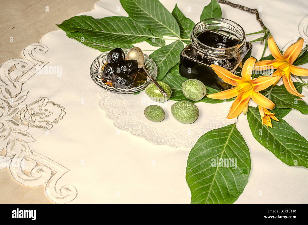 Crystal saucer and glass jar with nut jam near the green walnuts with leaves and orange lilies