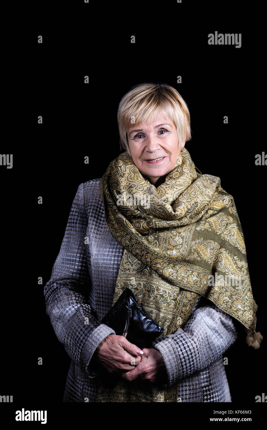 Portrait of an elderly woman in a draped coat with a thrown shawl on her shoulders and a clutch bag. Black background. Stock Photo