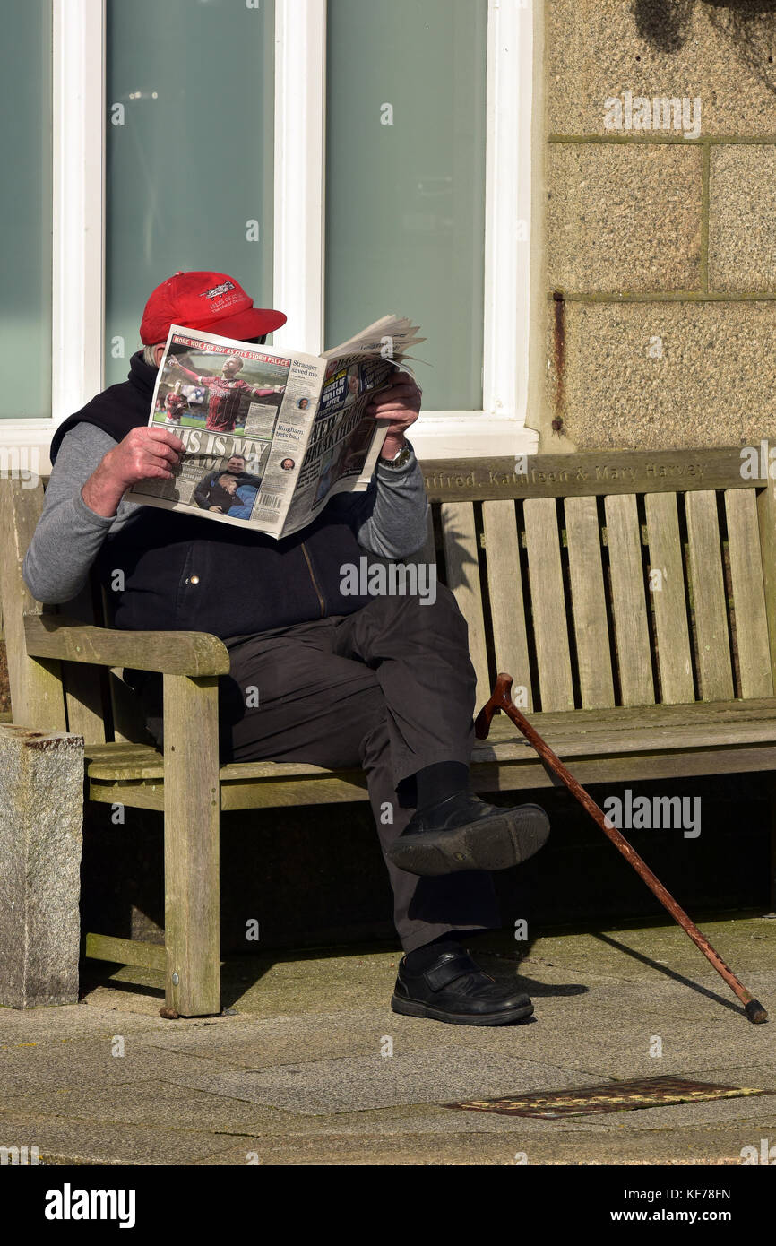 an elderly gentleman sitting on a bench reading a tabloid newspaper with his walking stick by his side in Saint Just, Cornwall, UK. Stock Photo