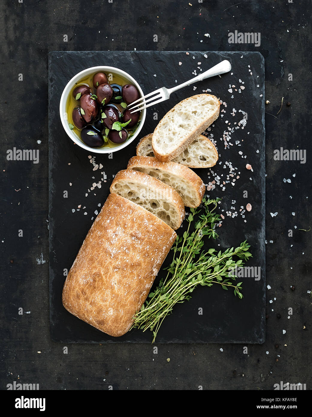 Italian ciabatta bread cut in slices on wooden chopping board with herbs, garlic and olives over dark grunge backdrop, top view Stock Photo
