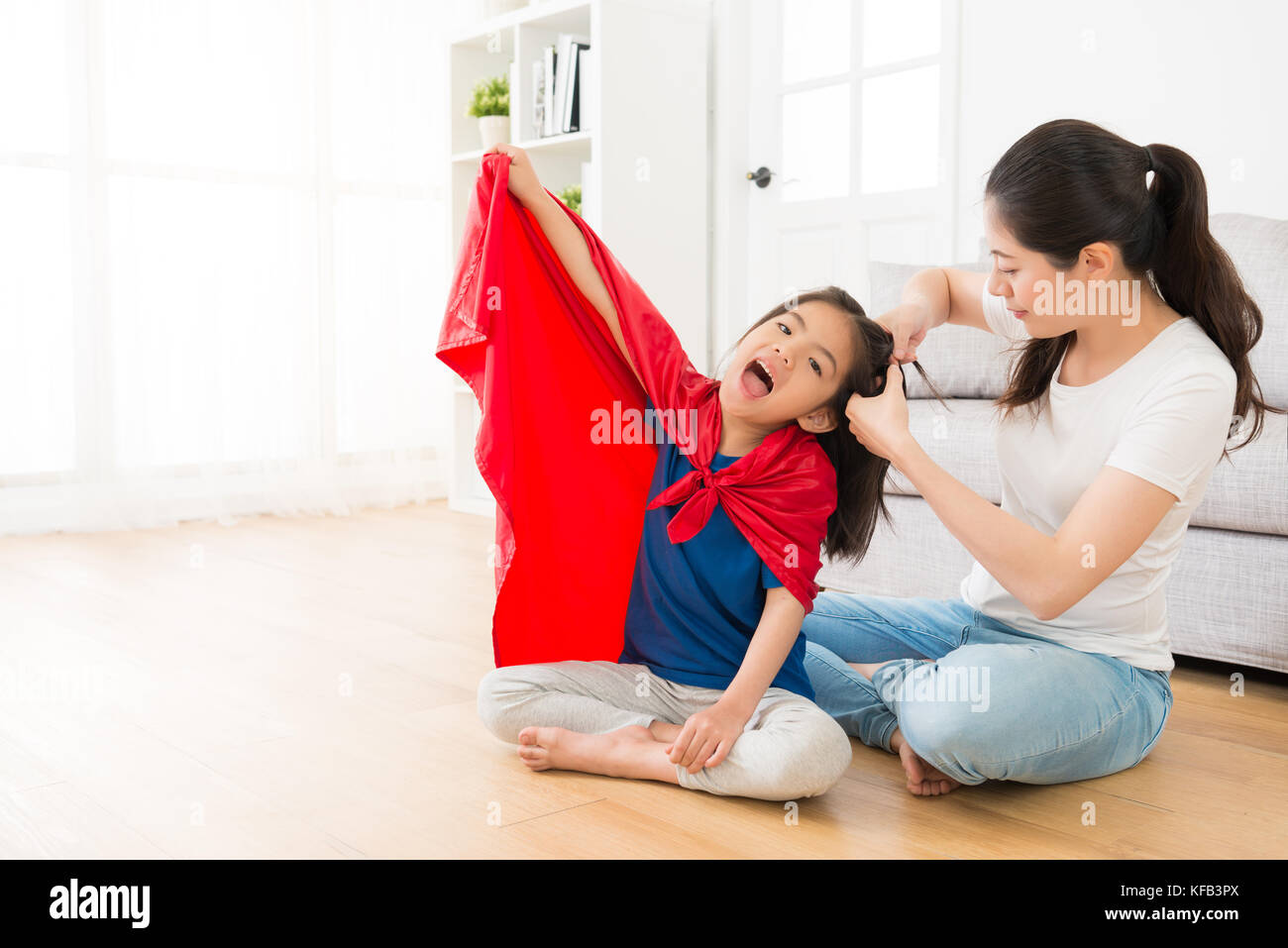 young smiling mother helping little girl tied hair in living room when daughter want to play as superhero at home with red cape. Stock Photo