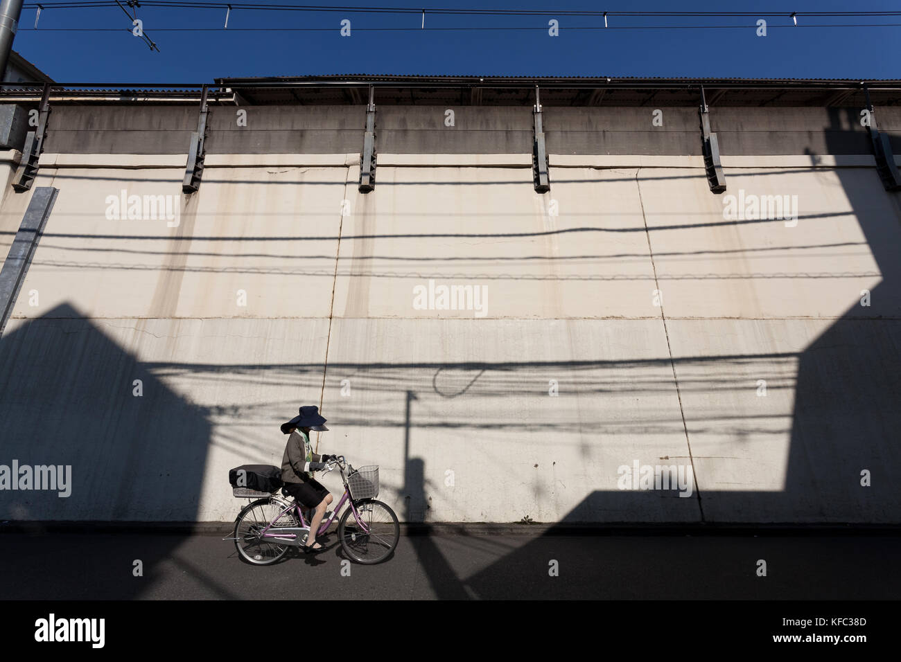 A Japanese woman in a large sun visor hat ride a bicycle by a wall near