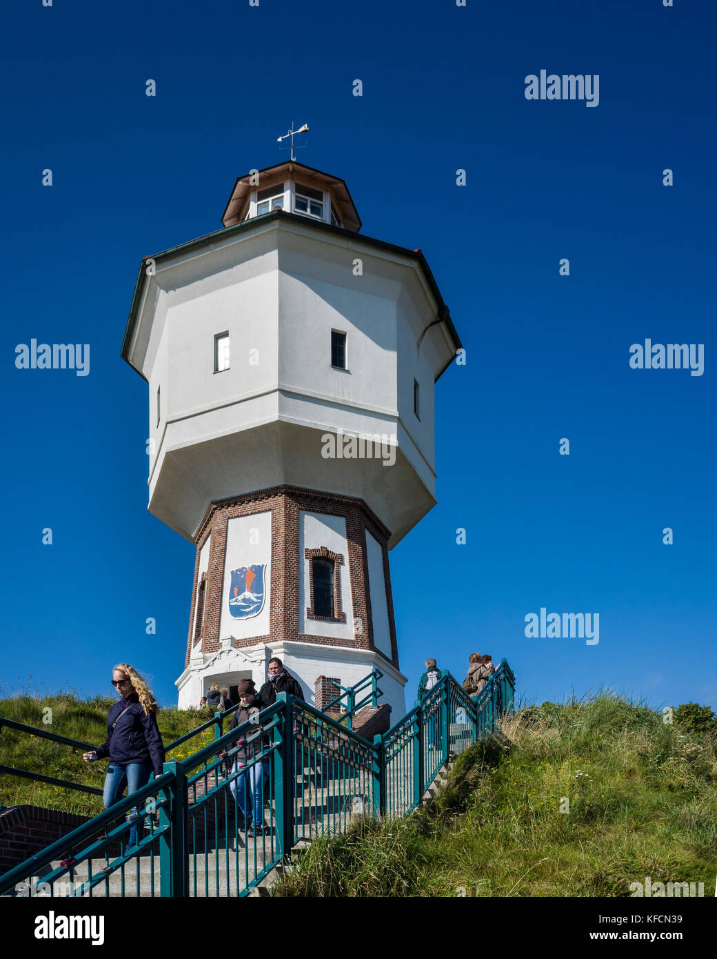 Wasserturm Langeoog. Germany Deutschland.  Holiday makers walking down the steps as the leave the water tower, which is one of Langeoog's main tourist attractions.  It's a sunny day making a nice blue sky to contrast the white of the water tower.  Previous visitors have left padlocks locked to the railings, which can be seen in the foreground.  Photographed with a Ricoh GRII camera. Stock Photo