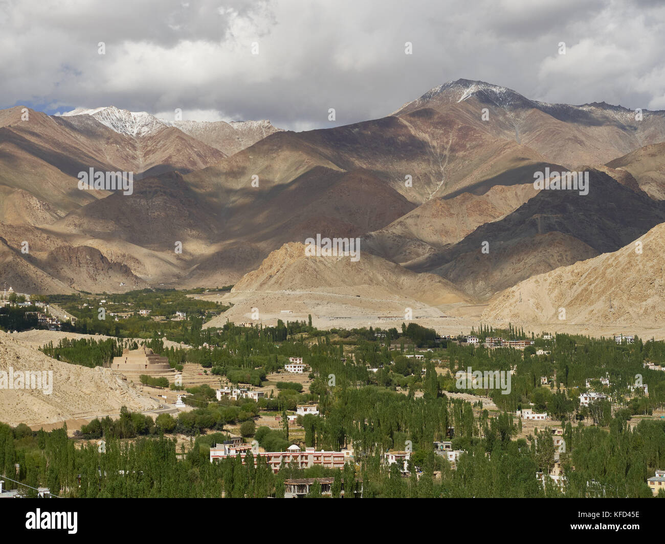 Huge mountain valley: brown mountains, shadows on the slopes, below in the wide part of the valley are many green trees and white houses, summer in th Stock Photo