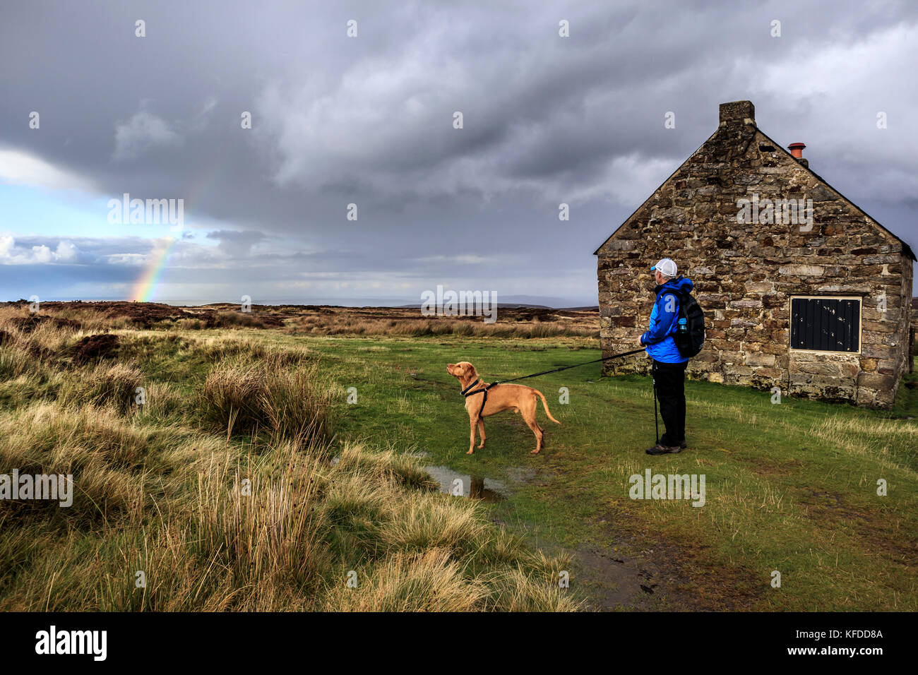 Walker looking at stormy skies and a rainbow with his dog by Trough house, an old shooting lodge on  Great Fryup Dale, North York Moors National Park Yorkshire England Stock Photo
