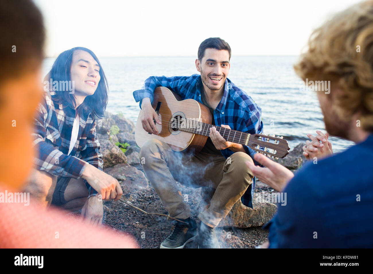 Happy guy with guitar and his friends spending evening around campfire by seaside Stock Photo