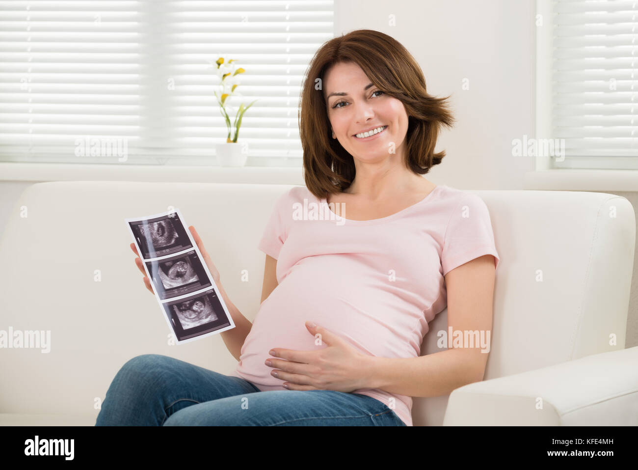 Happy Pregnant Woman Sitting On Sofa Holding Ultrasound Image Of Baby Stock Photo