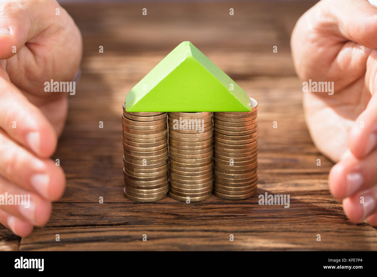 Close-up Of Hands Protecting The Coin House On Wooden Desk Stock Photo