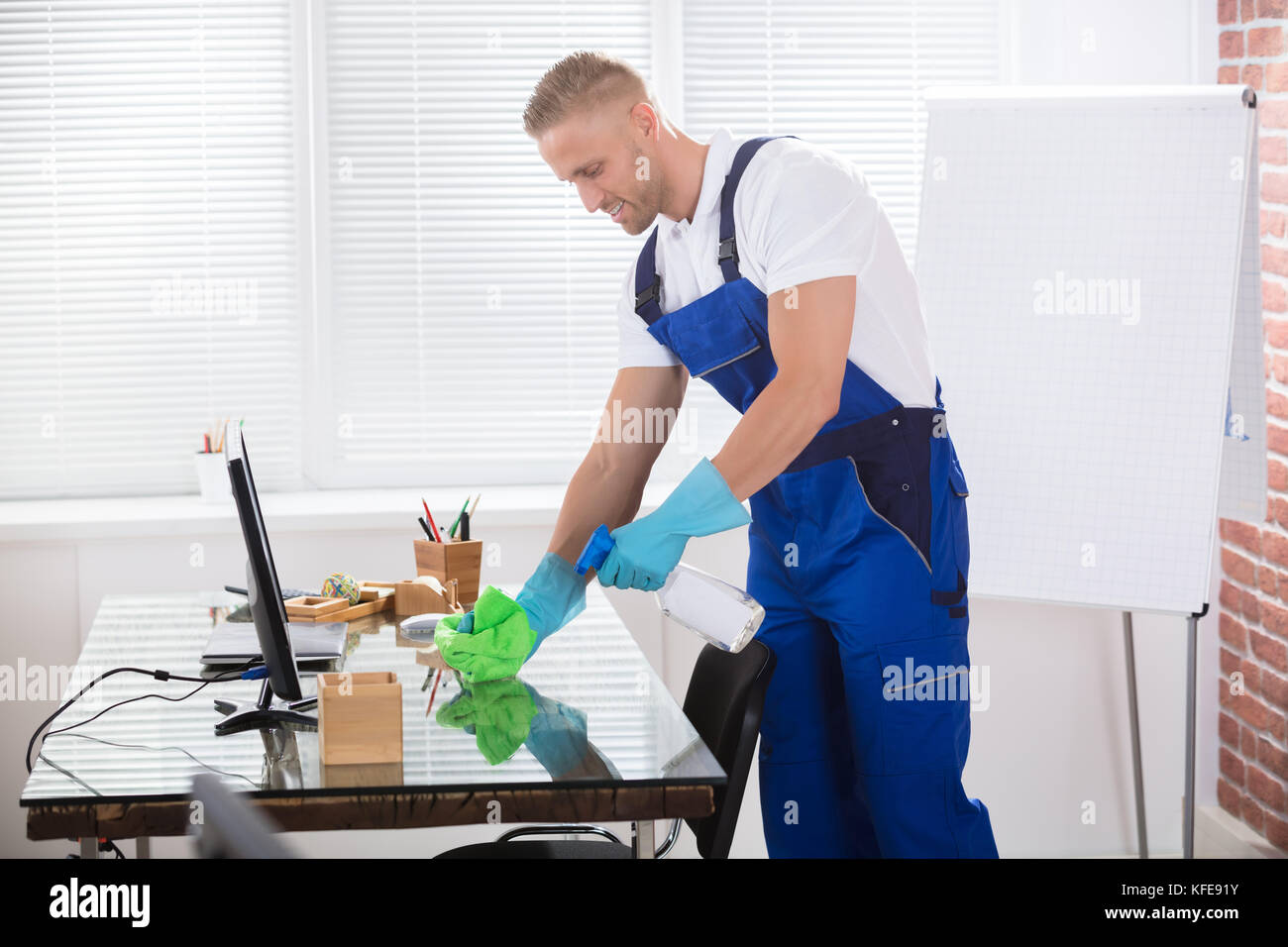 Portrait Of A Smiling Male Janitor Cleaning Desk With Cloth At Workplace Stock Photo