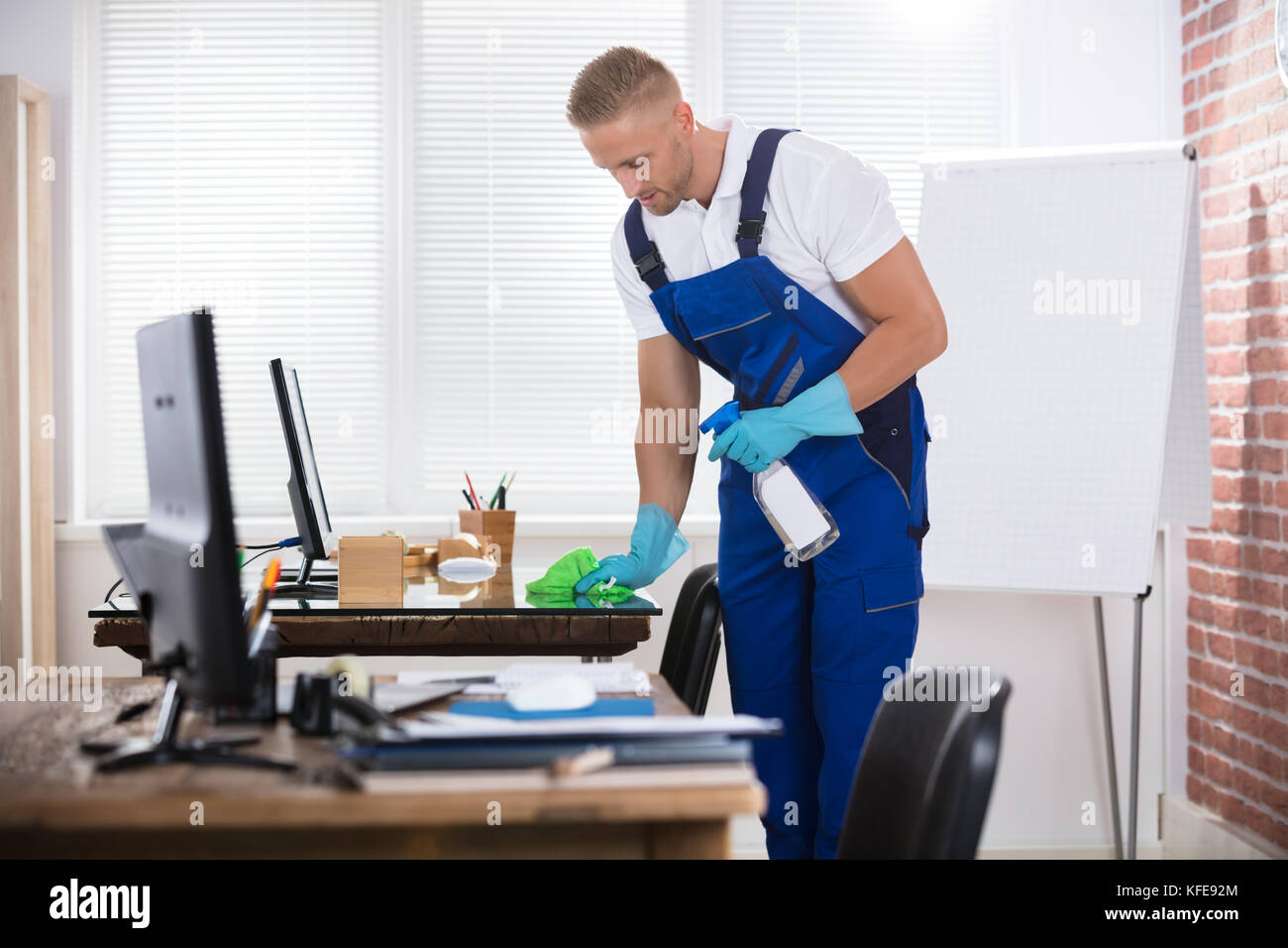 Portrait Of A Smiling Male Janitor Cleaning Desk With Cloth At Workplace Stock Photo
