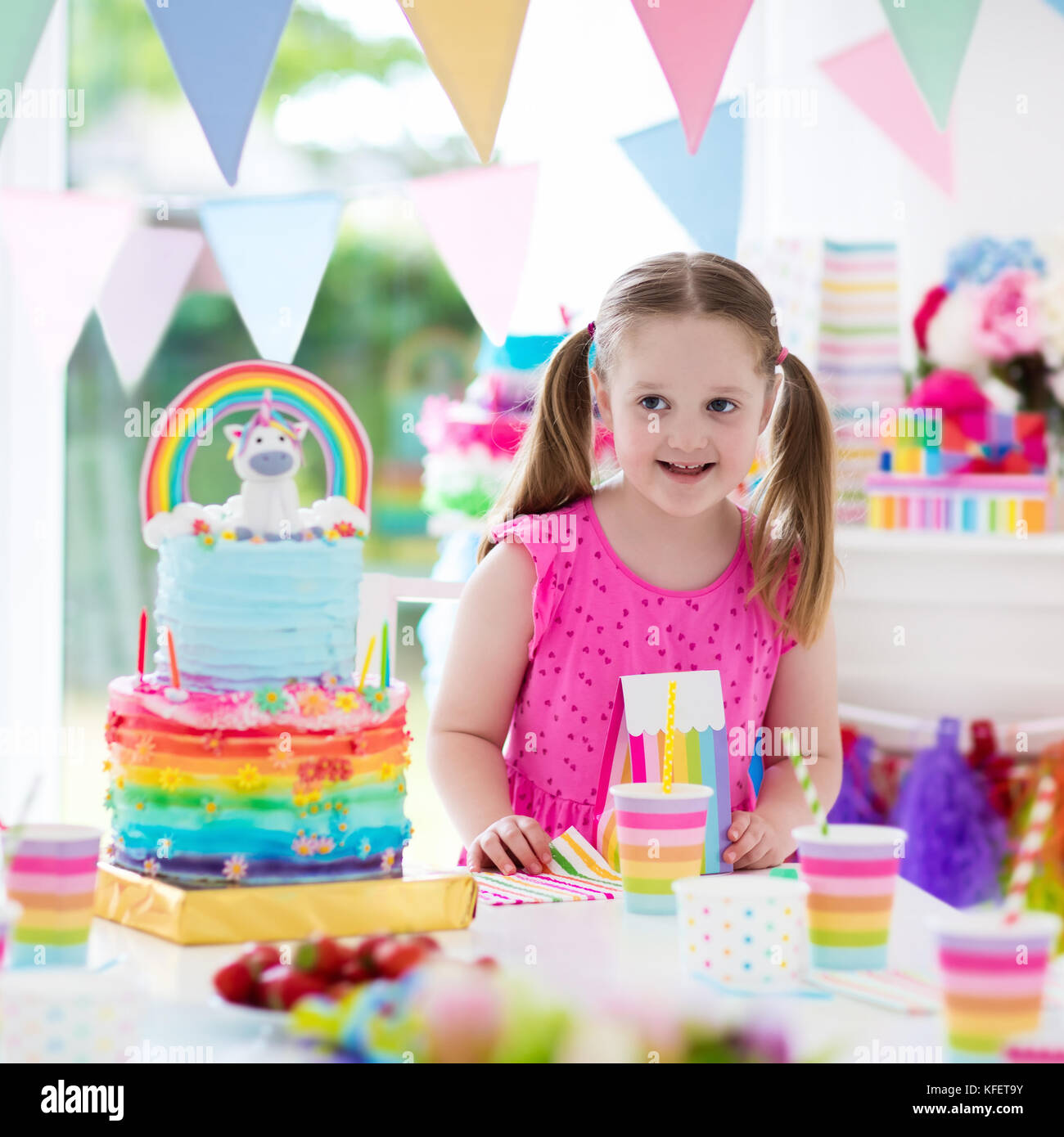 Kids birthday party with colorful pastel decoration and unicorn rainbow cake. Little girl with sweets, candy and fruit. Balloons and banner at festive Stock Photo