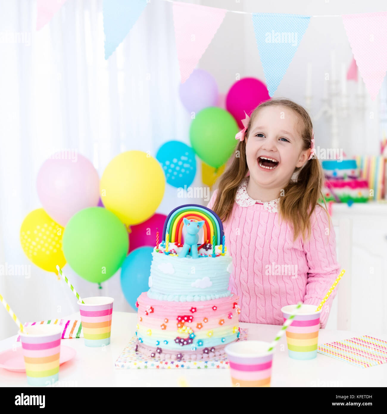 Kids birthday party with colorful pastel decoration and unicorn rainbow cake. Little girl with sweets, candy and fruit. Balloons and banner at festive Stock Photo