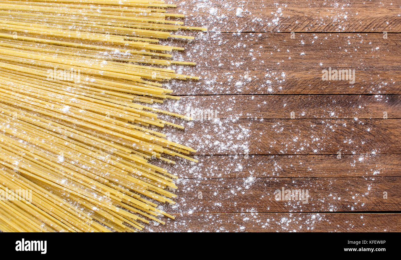 Spaghetti uncooked pasta on top of a light brown wooden surface, with lots of wheat flour sprinkled on top and around Stock Photo