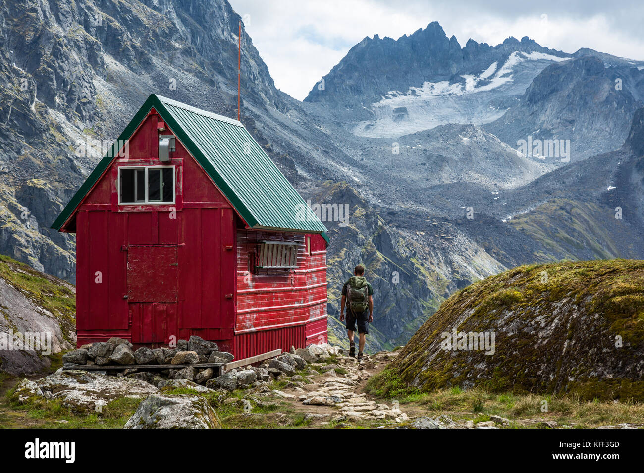 A man with a backpack out hiking walks up the trail to a red and green wilderness hut deep in the Talkeetna Mountains near Hatcher Pass, Alaska Stock Photo