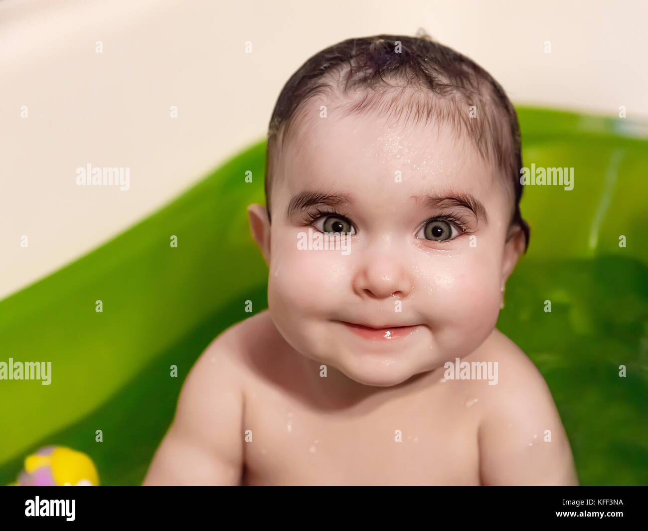 Smiling cute, pretty, happy baby girl bathing with a smile. Wet hair from the bath in a green bathtub. Nine months old / bathing smiling happy chubby Stock Photo
