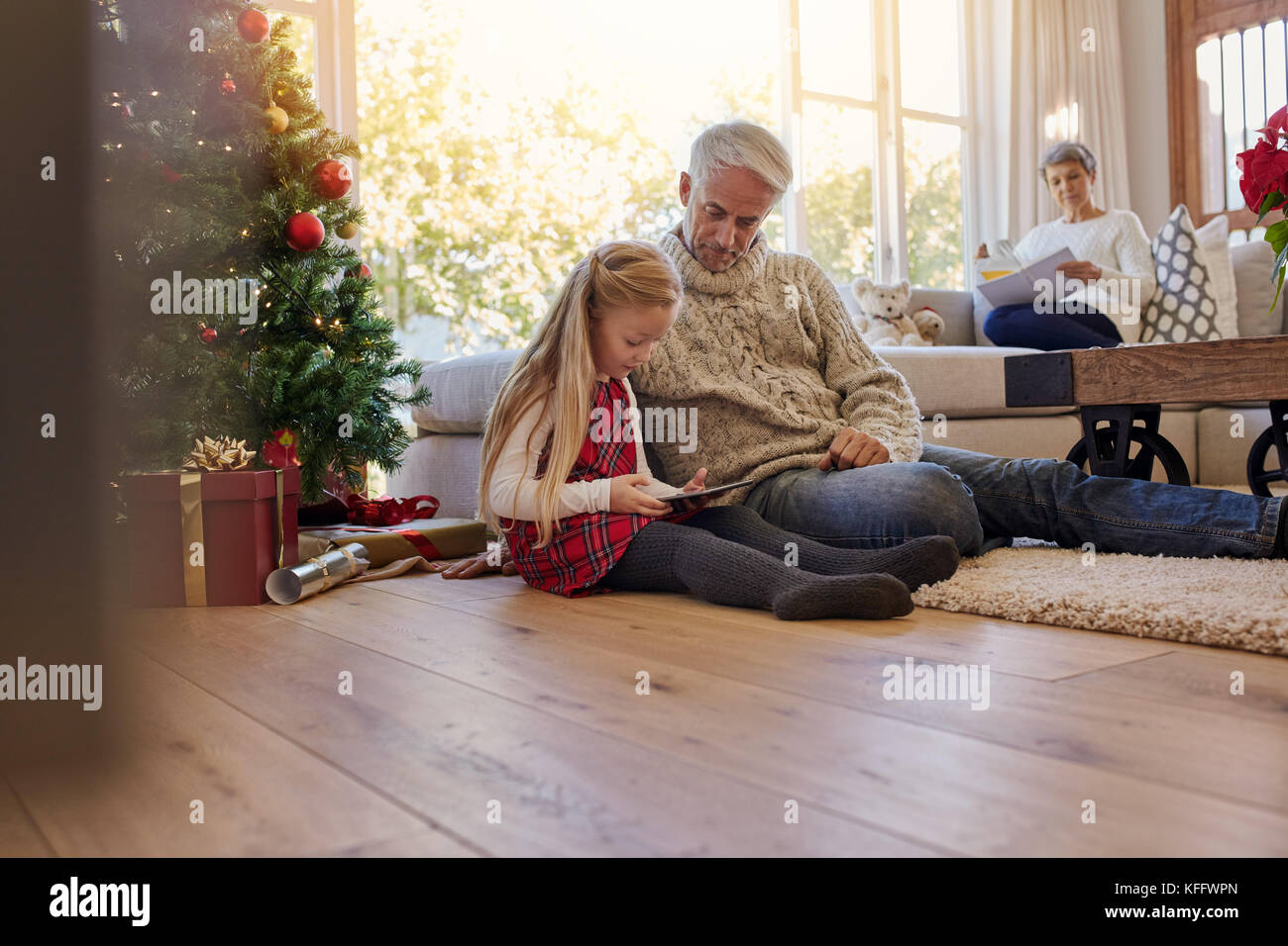 Little girl with grandfather sitting on floor and using digital tablet during Christmas, with grand mother sitting on sofa at the back. Little girl an Stock Photo